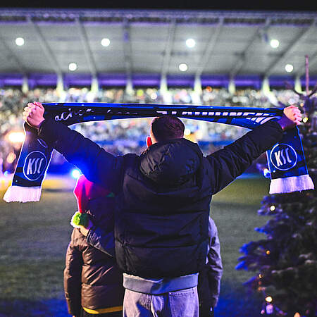 Kind hält einen KSC-Schal hoch, das Stadionsingen im Hintergrund