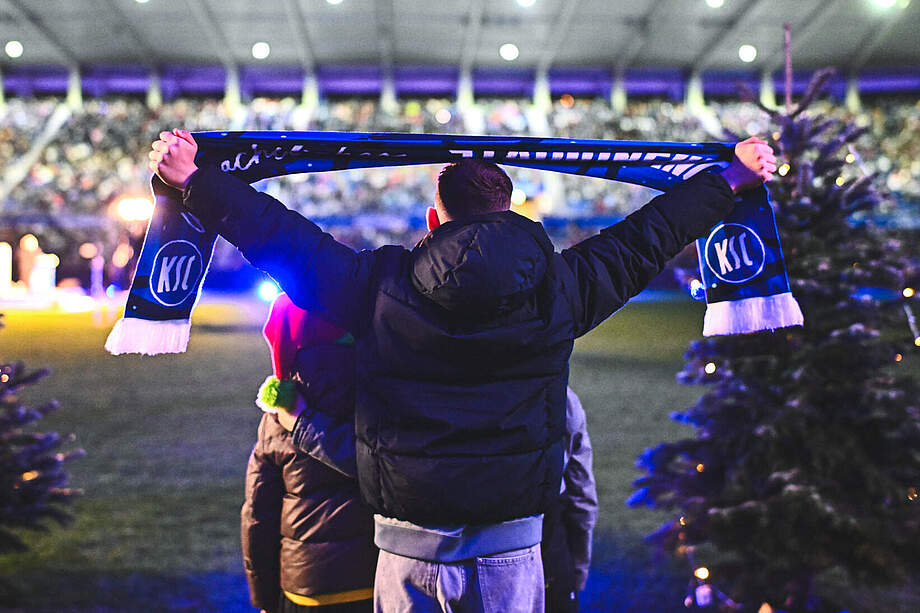 Kind hält einen KSC-Schal hoch, das Stadionsingen im Hintergrund