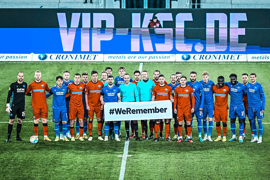 Die Spieler des KSC und des SC Paderborn halten ein Banner mit der Aufschrift "#WeRemeber" hoch. Das Bild stammt aus der Saison 2022/23.