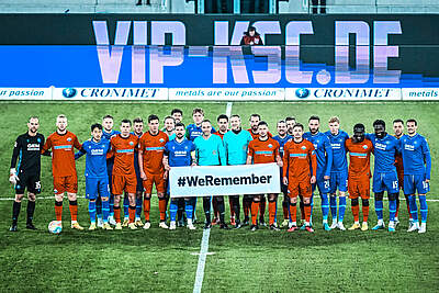 Die Spieler des KSC und des SC Paderborn halten ein Banner mit der Aufschrift "#WeRemeber" hoch. Das Bild stammt aus der Saison 2022/23.