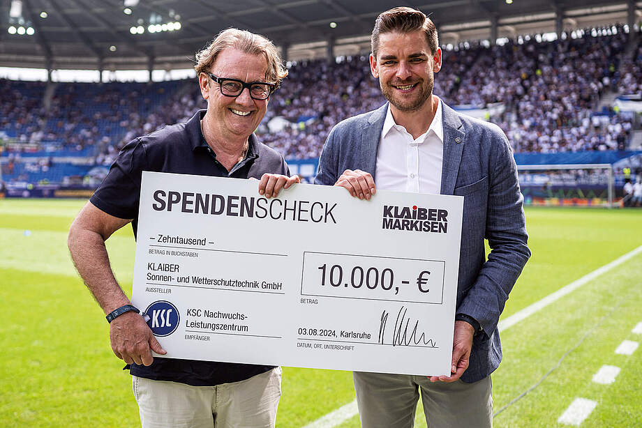 Ralf Klaiber und Michael Becker halten einen großen Scheck über 10.000 für die KSC aKAdemie in der Hand.